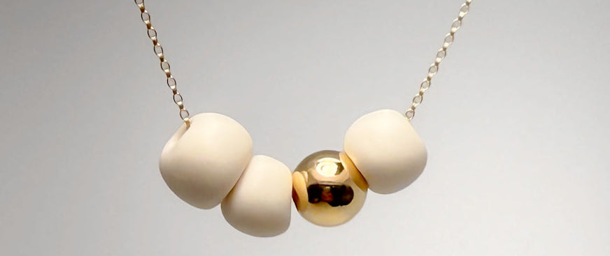 A close-up of a necklace with three matte whilte porcelain beads on a gold filled chain, and one polished gold bead, set against a neutral gray gradient background.