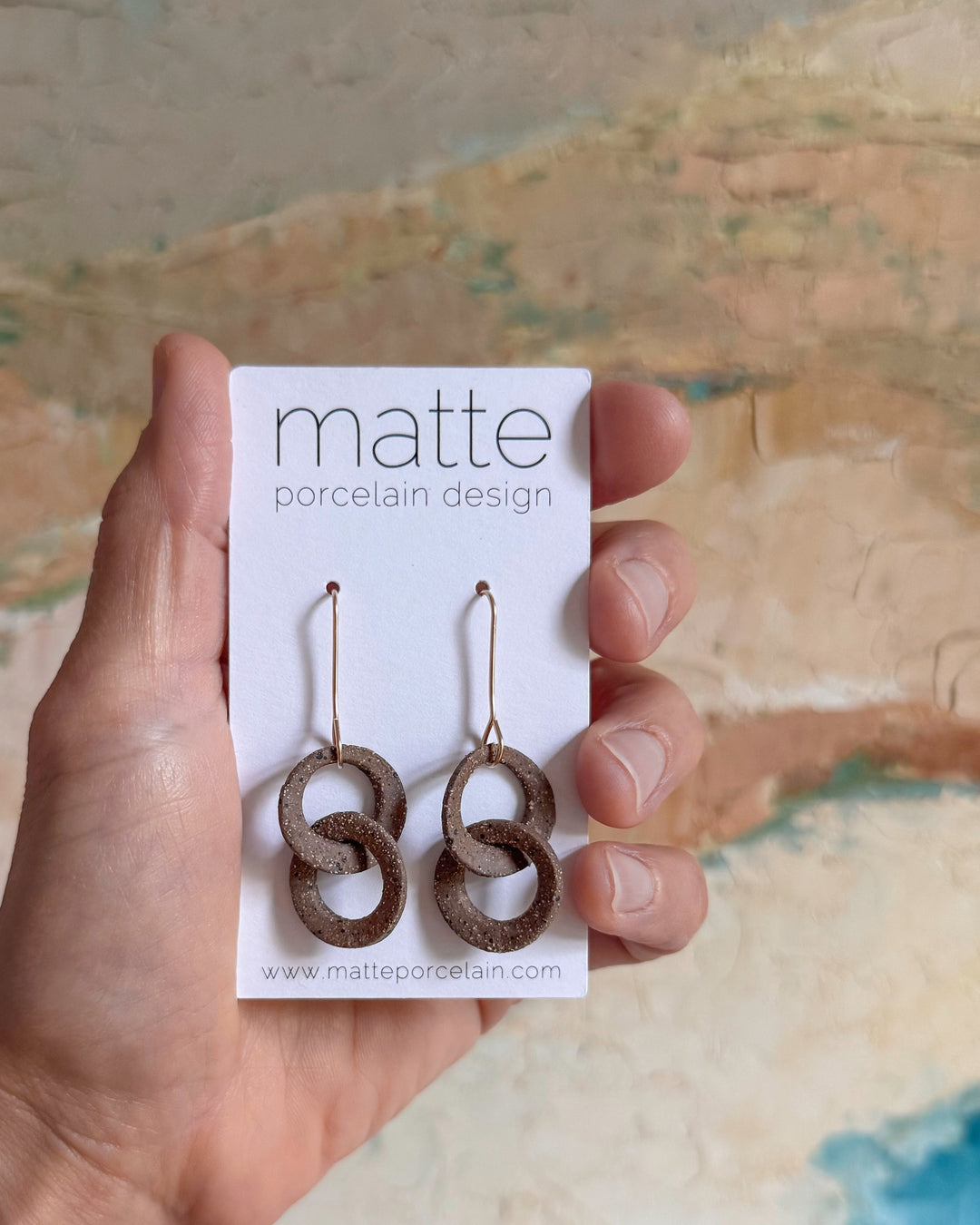 Hand holding a card with earrings labeled 'matte porcelain design' against a textured background.