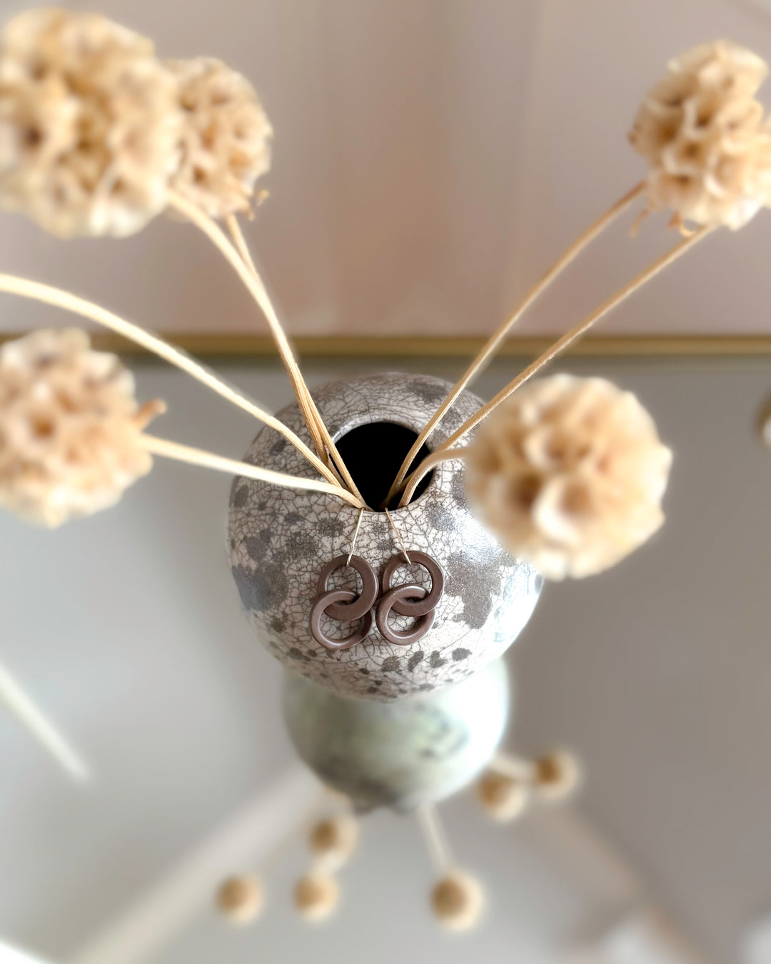 Porcelain earrings hang from decorative vase with dried flowers against a neutral background